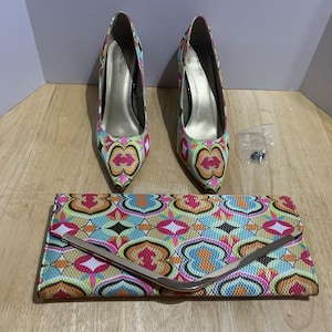 Puede incluir: Un conjunto a juego de zapatos de mujer y clutch. Los tacones de punta y el clutch presentan un colorido estampado geométrico en rosa, verde, azul y naranja. El clutch tiene un cierre plateado.