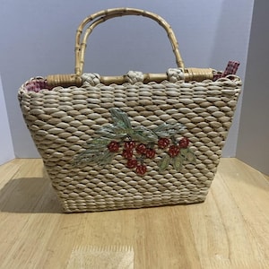 Puede incluir: Bolso de mano de paja tejida con asa de bambú. El bolso presenta un diseño bordado de bayas rojas y hojas verdes. El forro interior es un estampado de cuadros rojos y blancos. El bolso es de forma rectangular.