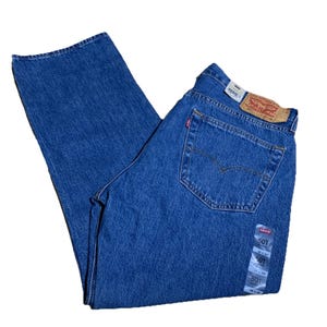 Puede incluir: Vaqueros azules de mezclilla con un diseño clásico de cinco bolsillos. Los vaqueros tienen un corte recto y una etiqueta roja de Levi's en el bolsillo trasero. Una etiqueta blanca con la talla 91x74 cm está adjunta. Los vaqueros están doblados.