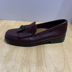 May include: A pair of burgundy leather loafers with a tassel detail. The shoes have a rounded toe and a low heel. The soles are black, and the shoes are sitting on a wooden surface.