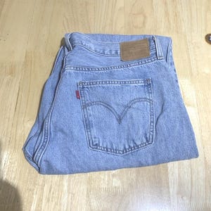 Puede incluir: Vaqueros de mezclilla azul claro con un diseño clásico de cinco bolsillos. Los vaqueros tienen un parche de cuero marrón en la parte trasera y una etiqueta roja en el bolsillo trasero. Los vaqueros están doblados sobre una superficie de madera.