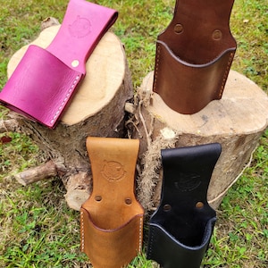 Könnte beinhalten: Vier Leder-Werkzeugtaschen in Pink, Braun, Hellbraun und Schwarz, jeweils mit einer Tasche und einer Schlaufe. Die Halter werden auf einem Baumstumpf und Gras präsentiert und zeigen die verschiedenen Farben und Designs.