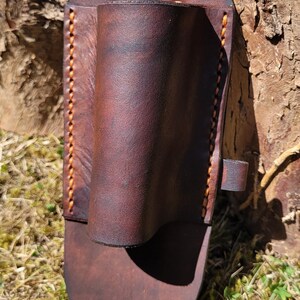 May include: A handcrafted, brown leather sheath with a cylindrical pocket and a flat base. The sheath features orange stitching along the edges and a small strap with a buckle. The leather has a rich, textured appearance.