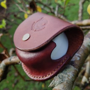 May include: A burgundy leather pouch with a white ball partially visible. The pouch is secured with a small, silver-coloured button. The words "Leather" are embossed on the pouch. The item is set against a blurred background of tree branches and foliage.