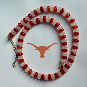 May include: A beaded necklace featuring alternating red and pink gemstones. The necklace has a gold-colored chain and clasp. A burnt orange longhorn logo is in the background.