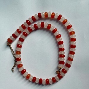 May include: A beaded necklace featuring alternating red and white beads. The necklace has a gold-colored chain and clasp. The beads are small and round, creating a delicate and elegant design. The necklace is displayed on a white surface.