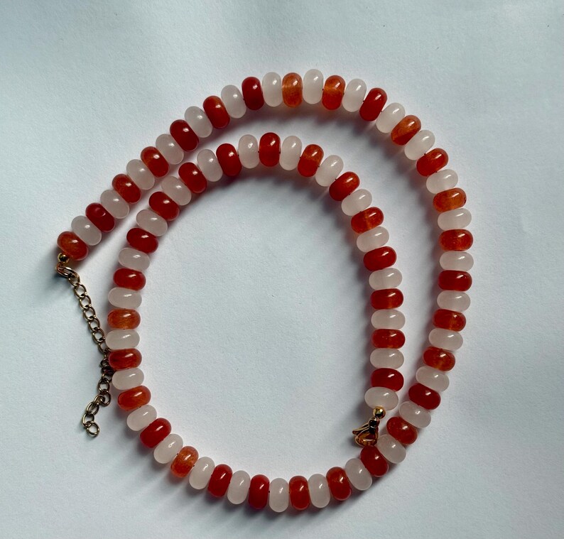 May include: A beaded necklace featuring alternating red and white beads. The necklace has a gold-colored chain and clasp. The beads are small and round, creating a delicate and elegant design. The necklace is displayed on a white surface.