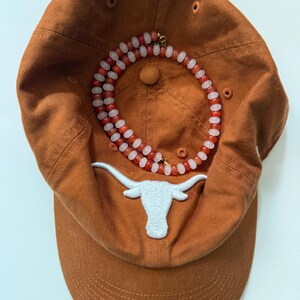 May include: An orange baseball cap with a white embroidered longhorn logo. A necklace with red and white beads is draped over the cap. The cap has a curved brim and a button on top.