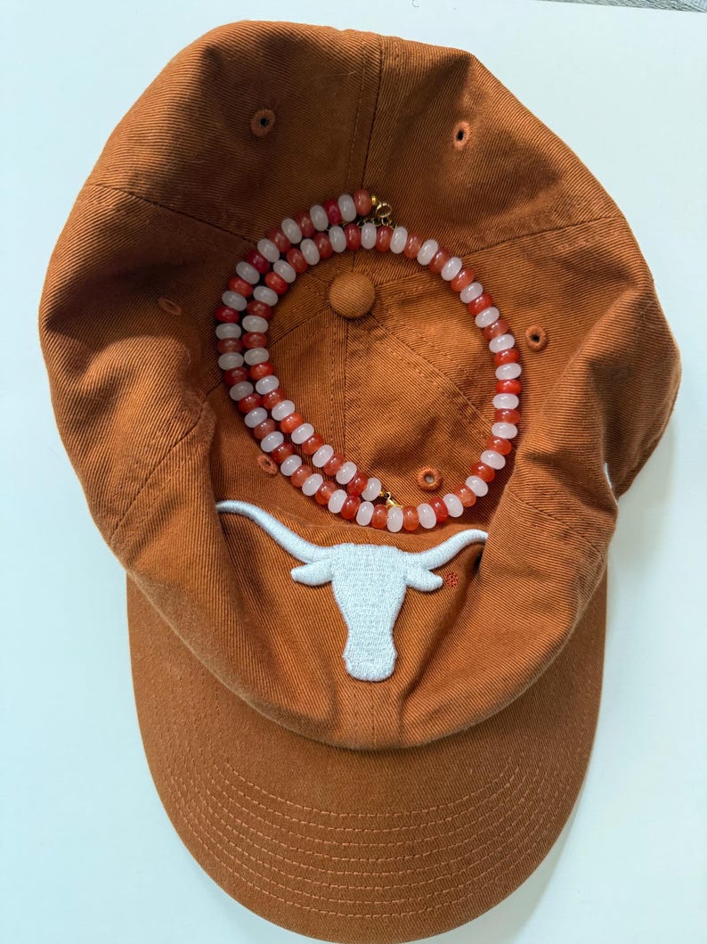 May include: An orange baseball cap with a white embroidered longhorn logo. A necklace with red and white beads is draped over the cap. The cap has a curved brim and a button on top.