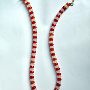 May include: A beaded necklace featuring alternating red and light pink beads. The necklace has a gold-colored clasp and chain. The beads are small and round, creating a delicate and colorful accessory. The necklace is displayed against a white background.