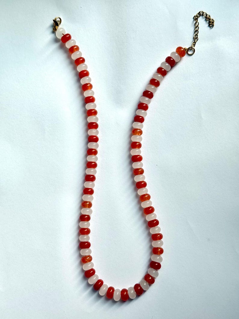 May include: A beaded necklace featuring alternating red and light pink beads. The necklace has a gold-colored clasp and chain. The beads are small and round, creating a delicate and colorful accessory. The necklace is displayed against a white background.