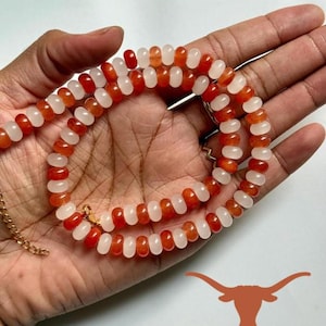 May include: A beaded necklace featuring alternating orange and white beads. The necklace is held in a hand, showcasing the color contrast and design. A gold-colored chain and clasp are visible.