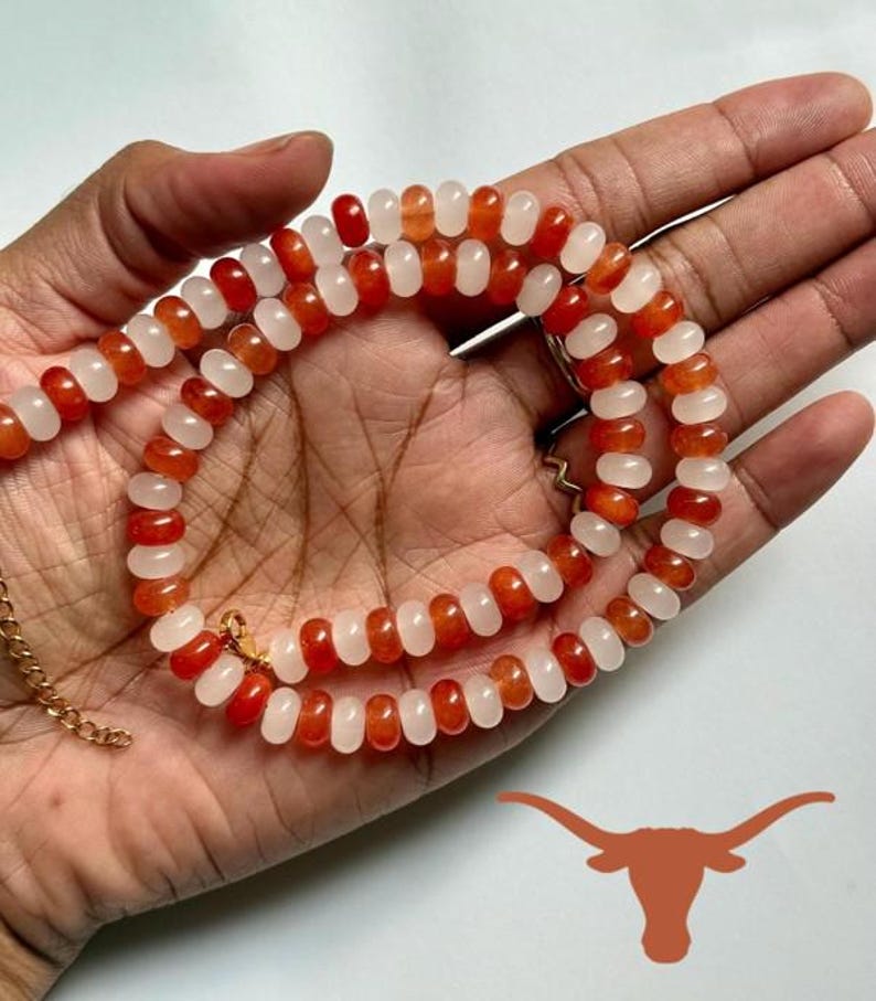 May include: A beaded necklace featuring alternating orange and white beads. The necklace is held in a hand, showcasing the color contrast and design. A gold-colored chain and clasp are visible.