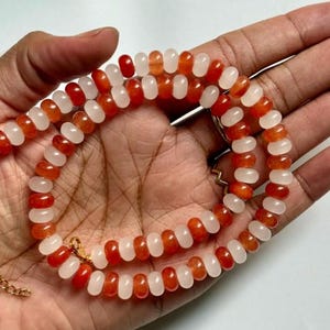 May include: A necklace composed of alternating orange and white beads. The beads are strung together with a gold-colored chain and clasp. The necklace is displayed on a hand.