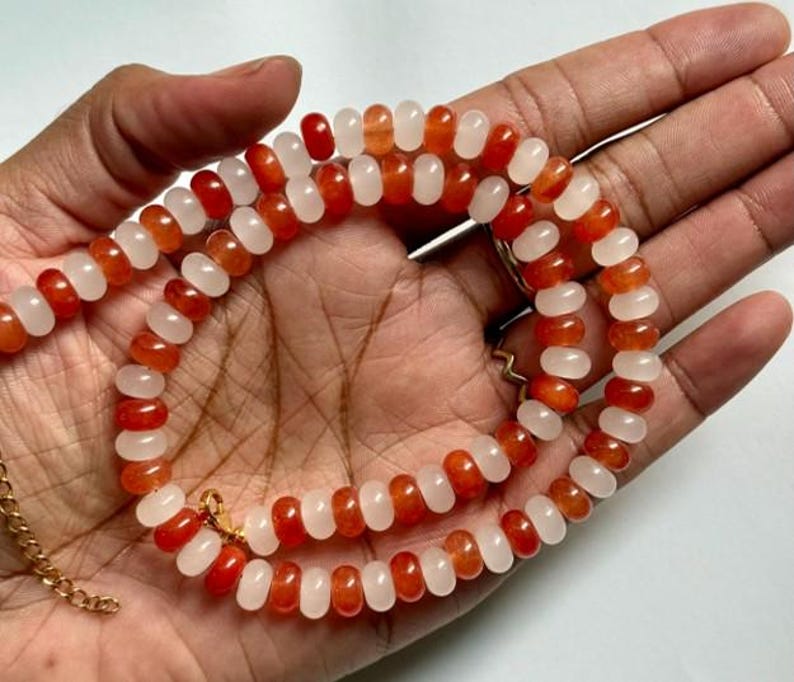 May include: A necklace composed of alternating orange and white beads. The beads are strung together with a gold-colored chain and clasp. The necklace is displayed on a hand.