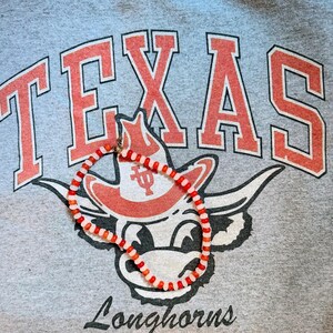 May include: A gray sweatshirt with the word "TEXAS" in red block letters and a Longhorns mascot graphic. A necklace with red and white beads is draped over the graphic.