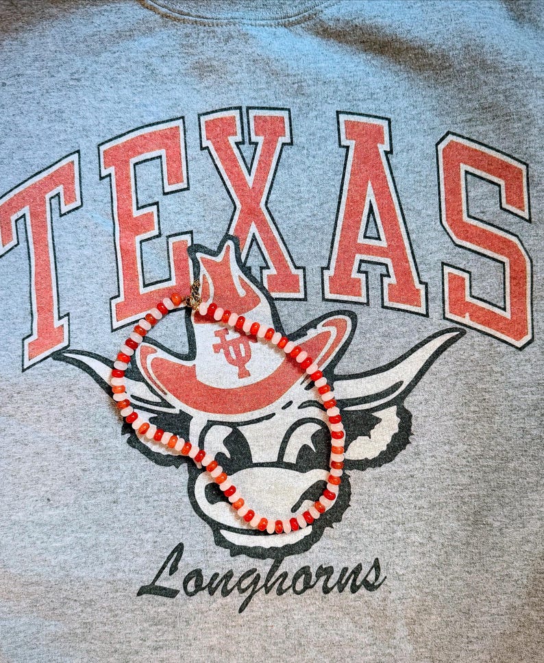 May include: A gray sweatshirt with the word "TEXAS" in red block letters and a Longhorns mascot graphic. A necklace with red and white beads is draped over the graphic.