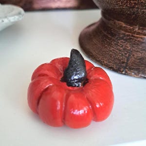 Peut inclure: Une petite figurine de citrouille rouge avec une tige noire. La citrouille décorative est posée sur une surface blanche, avec un vase marron en arrière-plan. La citrouille est une décoration sur le thème de l'automne.