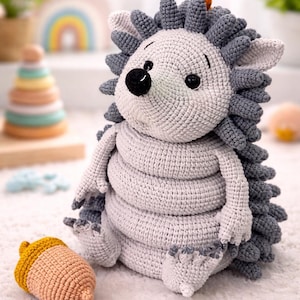 Könnte beinhalten: Ein handgehäkeltes Igel-Spielzeug in Hellgrau und Dunkelgrau. Der Igel hat einen runden Körper, ein lächelndes Gesicht und eine Reihe von Stacheln. Ein eichelförmiges Spielzeug in Gelb und Pfirsich liegt neben dem Igel. Das Spielzeug ist für Babys.