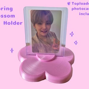 May include: A pink, flower-shaped card holder with a clear plastic top. The holder displays a photo card. Text on the image reads "Spring Blossom Card Holder". The background is light pink with purple star accents.