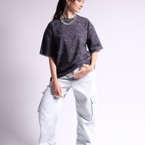 Pode incluir: Uma t-shirt de manga curta cinzento-carvão com lavagem ácida combinada com calças cargo de ganga de lavagem clara com bolsos laterais. O conjunto é complementado por um colar de cor prateada em camadas e ténis desportivos brancos.
