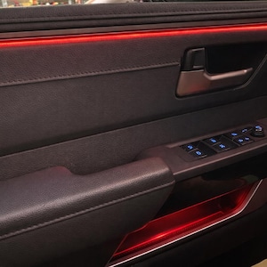 May include: Car door interior panel in dark grey with a leather-like texture. It has a chrome door handle, power window controls with blue lighting, and red accent lighting along the top and bottom edges.