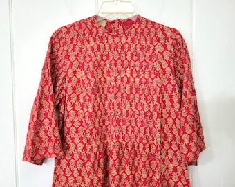 May include: A red dress with a floral pattern in tan and black. The dress has a high collar, long sleeves, and pleats down the front. The dress is hanging on a white hanger.