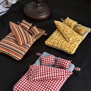 May include: Three miniature bed sets featuring rectangular mattresses, pillows, and blankets. One set has orange, black, and white stripes. Another set features a yellow and white floral plaid pattern. The third set displays a red and white gingham check.