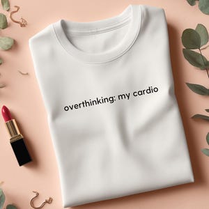 Könnte beinhalten: Weißes Sweatshirt mit Rundhalsausschnitt und dem Text "overthinking: my cardio" in Schwarz. Das Sweatshirt wird auf einer pfirsichfarbenen Oberfläche mit rotem Lippenstift, goldenen Ohrringen und Grünzeug präsentiert.