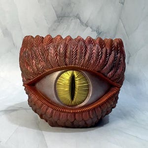 May include: A cylindrical planter featuring a textured exterior of overlapping copper-colored feathers. Centered on the front is a large, realistic eye with a metallic silver iris and a vertical, bright gold pupil, creating a striking focal point.