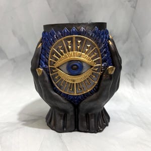 May include: A decorative vessel featuring two dark gray hands cupping a central circular emblem. The emblem displays a gold-toned eye with a blue iris, surrounded by radiating gold rays and a textured blue border with pointed, petal-like details.