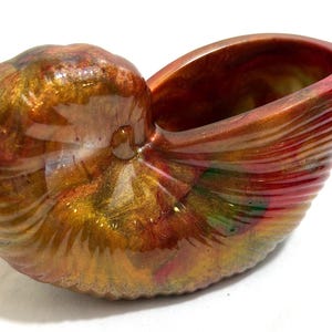 May include: A decorative nautilus shell-shaped container. The shell is a mix of colors, including gold, red, and green, with a glossy finish. The container has a smooth interior and a textured exterior, resembling the natural shell.