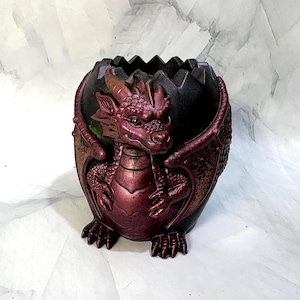 May include: A dark-colored, egg-shaped container featuring a sculpted, metallic burgundy dragon emerging from the front. The dragon has detailed scales, wings, and horns, while the top edge of the container is jagged, resembling a cracked eggshell.
