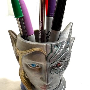 May include: A unique pen holder featuring a split design: one side depicts a human face with blonde hair and blue eyes, the other a robotic face. The holder contains pens and markers in various colors, including red, green, and purple.