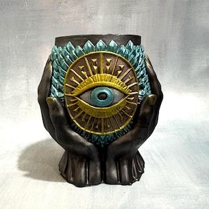 May include: A dark-toned sculptural vessel shaped like two cupped hands holding a central circular emblem. The emblem features a prominent blue eye surrounded by gold rays and a textured teal-colored feather-like pattern encircling the rim.