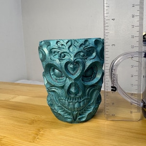 May include: A teal-colored, decorative skull-shaped container. The container has intricate carved details, including heart shapes and floral patterns. The container is approximately 3 inches tall.