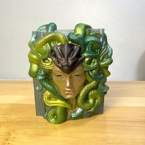 May include: A decorative, square-shaped Medusa head sculpture. The face is gold, with a dark brown crown. The snakes are green and yellow. The sculpture is on a wooden surface.