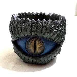 May include: A decorative, dark green planter with a detailed feather design around the rim and base. The planter features a large, blue eye with a yellow and brown iris in the center. The planter is likely made of resin or a similar material.