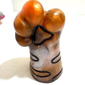 May include: A unique, abstract art piece with a bulbous top and a cylindrical base. The sculpture features a blend of metallic orange and brown hues, accented with black outlines. The piece has a glossy finish.