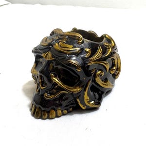 May include: A decorative skull-shaped container in black with gold accents. The skull features ornate, swirling gold patterns and a hollowed-out top. The container is likely used for holding small items or as a unique decorative piece.