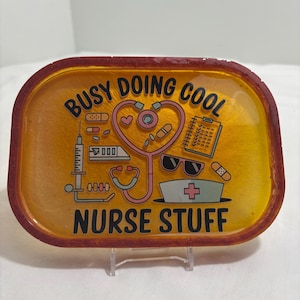 May include: A rectangular tray with a yellow and orange design. The tray has a red border and features medical-themed graphics, including a stethoscope, syringe, and the text "BUSY DOING COOL NURSE STUFF."