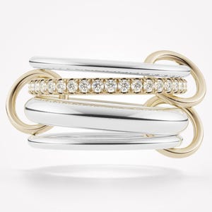 Pode incluir: Anel de várias faixas com uma combinação de tons prateados e dourados. O anel apresenta uma faixa central adornada com pequenas pedras brilhantes. O design incorpora elementos interligados, criando um visual moderno e sofisticado.