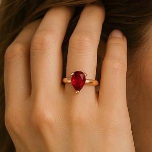Op de afbeelding: Een gouden ring met een enkele peervormige rode edelsteen in een zetting met drie pootjes. De ringband is glad en gepolijst, met een strak ontwerp dat de levendige kleur en de druppelvormige slijpvorm van de centrale steen benadrukt.