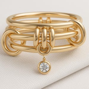 May include: A gold ring with a distinctive design. The ring has multiple interconnected bands and a dangling, round, clear gemstone. The ring is displayed on a white surface, highlighting its intricate details and polished finish.