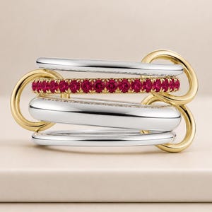 Op de afbeelding: Een meerbandige ring met zilver- en goudtinten. Eén band is versierd met een rij kleine, felrode edelstenen. De ring is verbonden door goudkleurige lussen, wat een uniek en stijlvol ontwerp creëert.