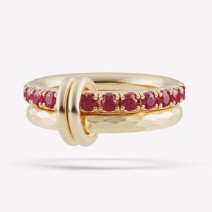 May include: A gold ring set consisting of two interlocking bands. One band features a row of round, vibrant red gemstones set in gold prongs. The second band has a textured, hammered finish. Both bands are connected by a smooth gold loop.