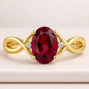 Puede incluir: Un anillo de oro con una gran piedra preciosa de color rojo intenso y corte ovalado en el centro, flanqueada por dos pequeñas piedras redondas y transparentes. La banda tiene un diseño trenzado y cruzado que se une al engaste central.