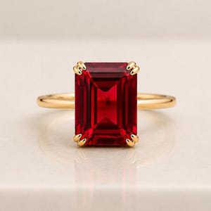 May include: A gold ring featuring a large, rectangular emerald-cut deep red gemstone set in a four-prong setting. The band is smooth and polished, showcasing a simple design that highlights the vibrant color and faceted clarity of the center stone.