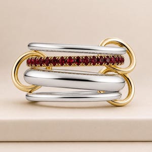 May include: A multi-band ring featuring silver and gold tones. The ring has three silver bands, one gold band, and a gold band with small red gemstones. The bands are interlocked, creating a unique design.