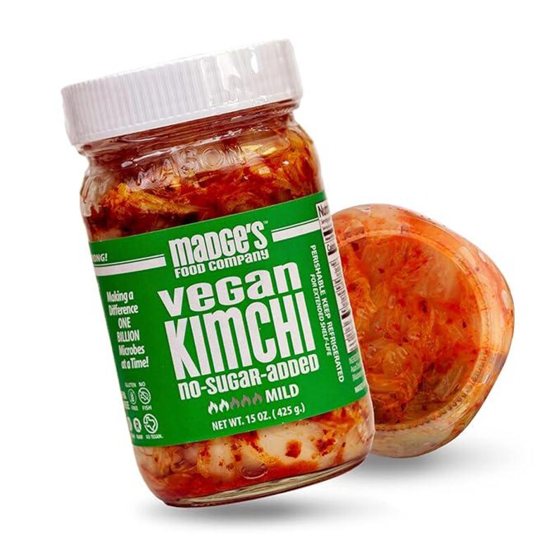 May include: A jar of Madge's Food Company Vegan Kimchi with a green label. The label reads "Vegan Kimchi, no-sugar-added, mild". The jar is filled with red kimchi and has a white lid. A container of kimchi is next to the jar.